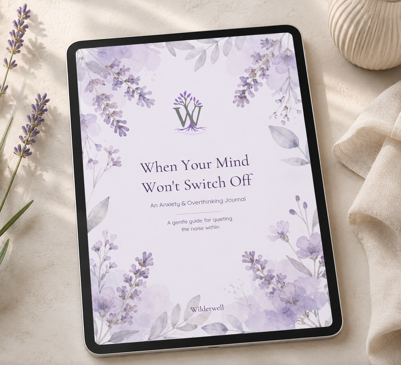 A tablet displaying a lavender watercolor journal cover with the title 'When Your Mind Won't Switch Off' and the brand name 'Wilderwell', styled on a light beige surface with lavender sprigs and a cream textured bowl.