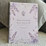 A spiral-bound journal titled 'When Your Mind Won't Switch Off' by Wilderwell, featuring a lavender and gray floral watercolor design on a pale background, resting on a textured blanket with a gray pillow behind it.