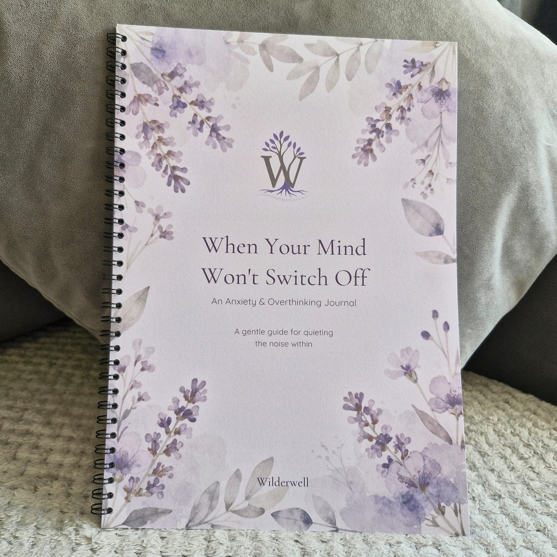 A spiral-bound journal titled 'When Your Mind Won't Switch Off' by Wilderwell, featuring a lavender and gray floral watercolor design on a pale background, resting on a textured blanket with a gray pillow behind it.