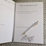 An open spiral-bound journal or planner spread on a textured gray blanket, showing printed prompts such as 'What's Going Well in My Life?' and a glittery pastel pen placed diagonally across the pages.