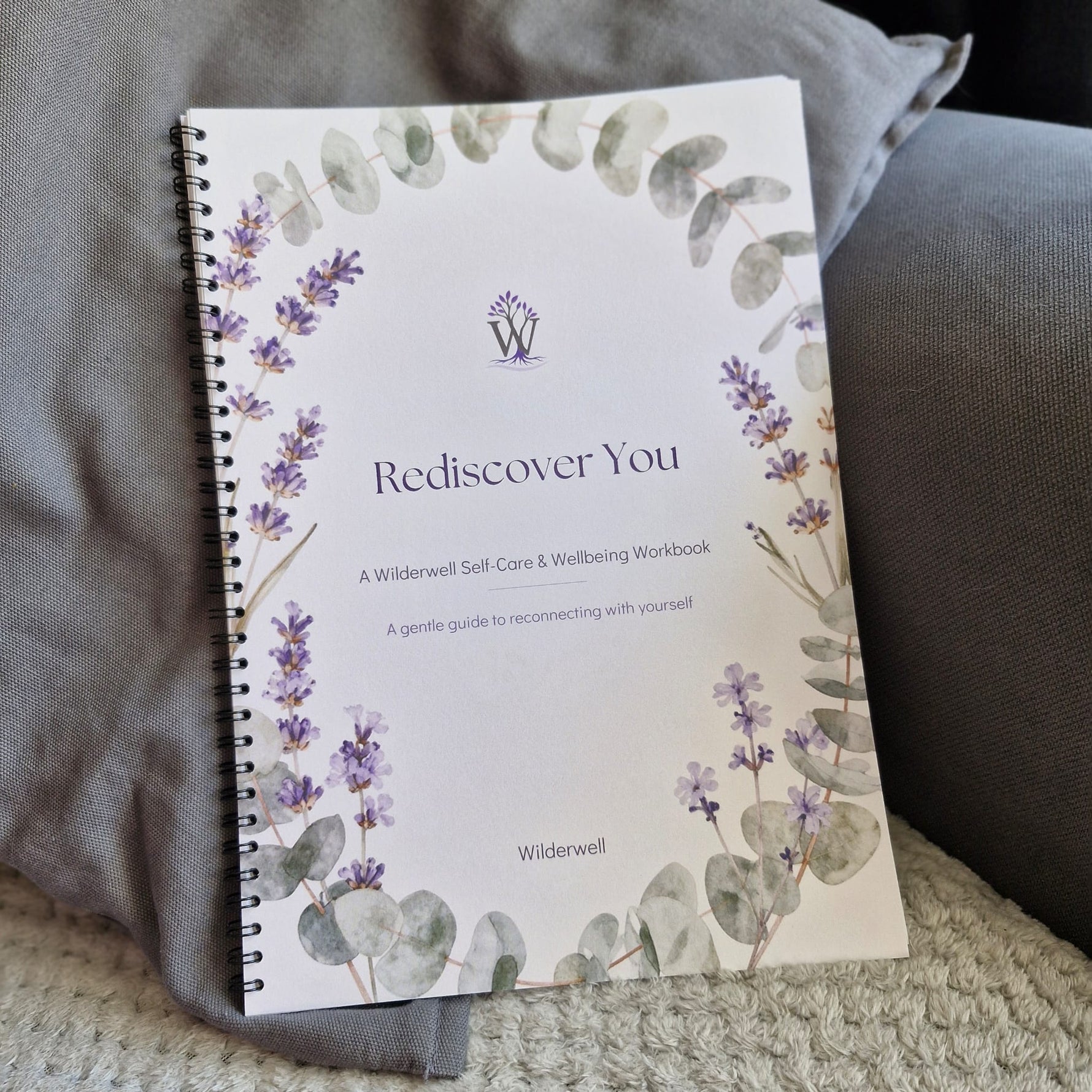 A spiral-bound workbook titled "Rediscover You" by Wilderwell, placed on a gray cushion with a textured blanket underneath. The cover features watercolor illustrations of lavender and eucalyptus.